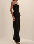 A woman modeling a black strapless top and matching wide-leg trousers set, paired with black heeled sandals, in a minimalist cream studio setting.