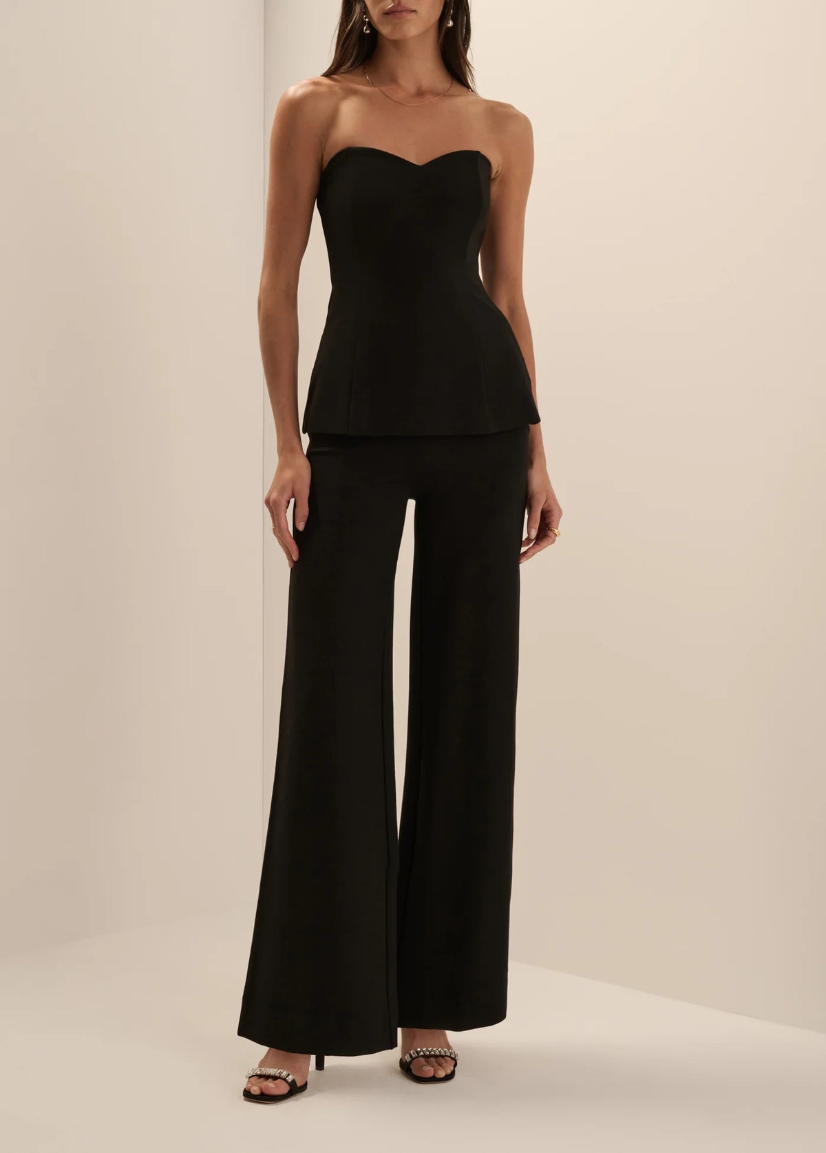 A woman modeling a black strapless peplum top and matching wide-leg pants set against a plain beige studio background.