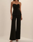 A woman modeling a black strapless peplum top and matching wide-leg pants set against a plain beige studio background.