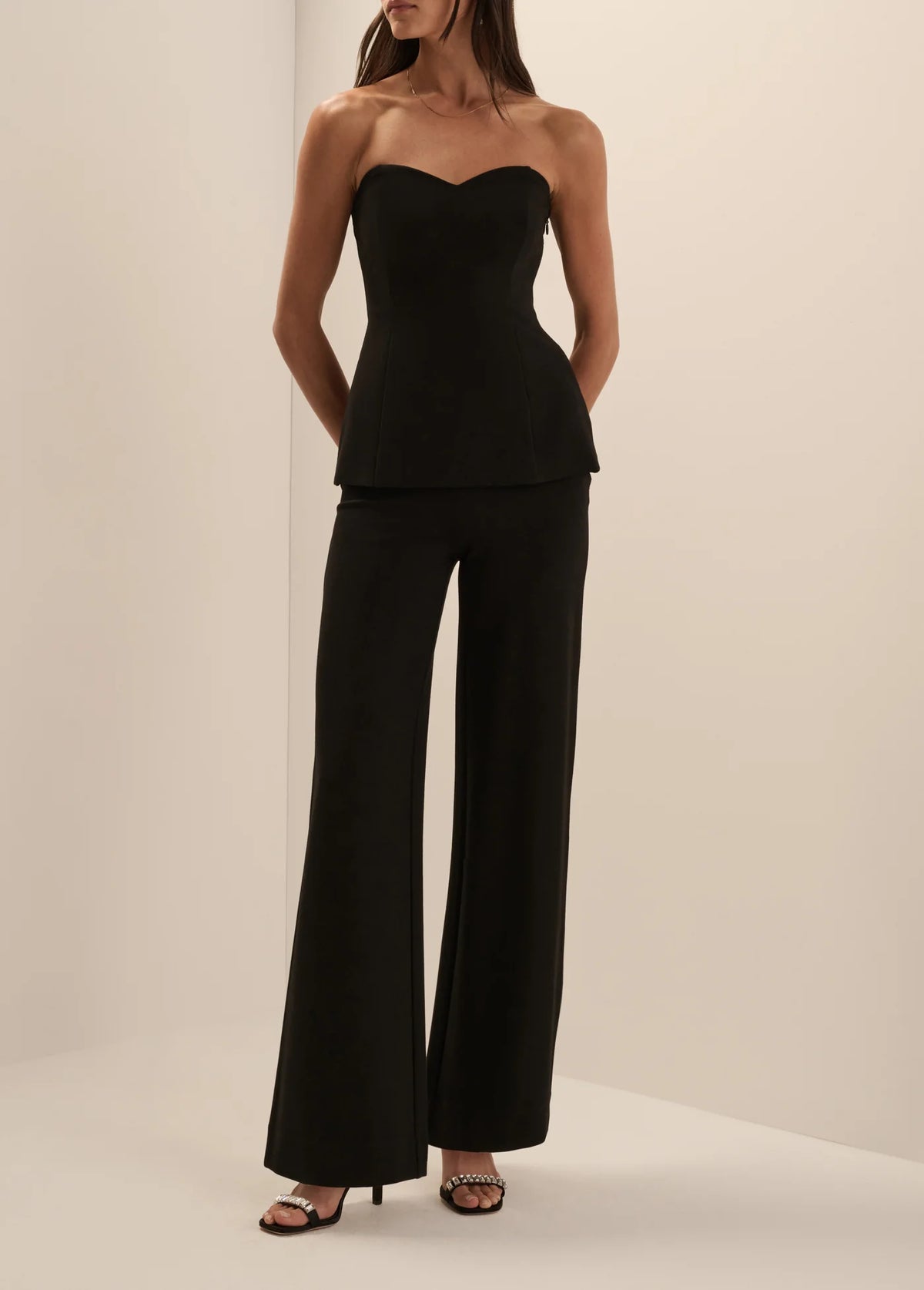 A woman modeling a black strapless tube top and matching wide-leg pants set, standing against a plain warm off-white studio background.