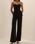 A woman modeling a black strapless tube top and matching wide-leg pants set, standing against a plain warm off-white studio background.