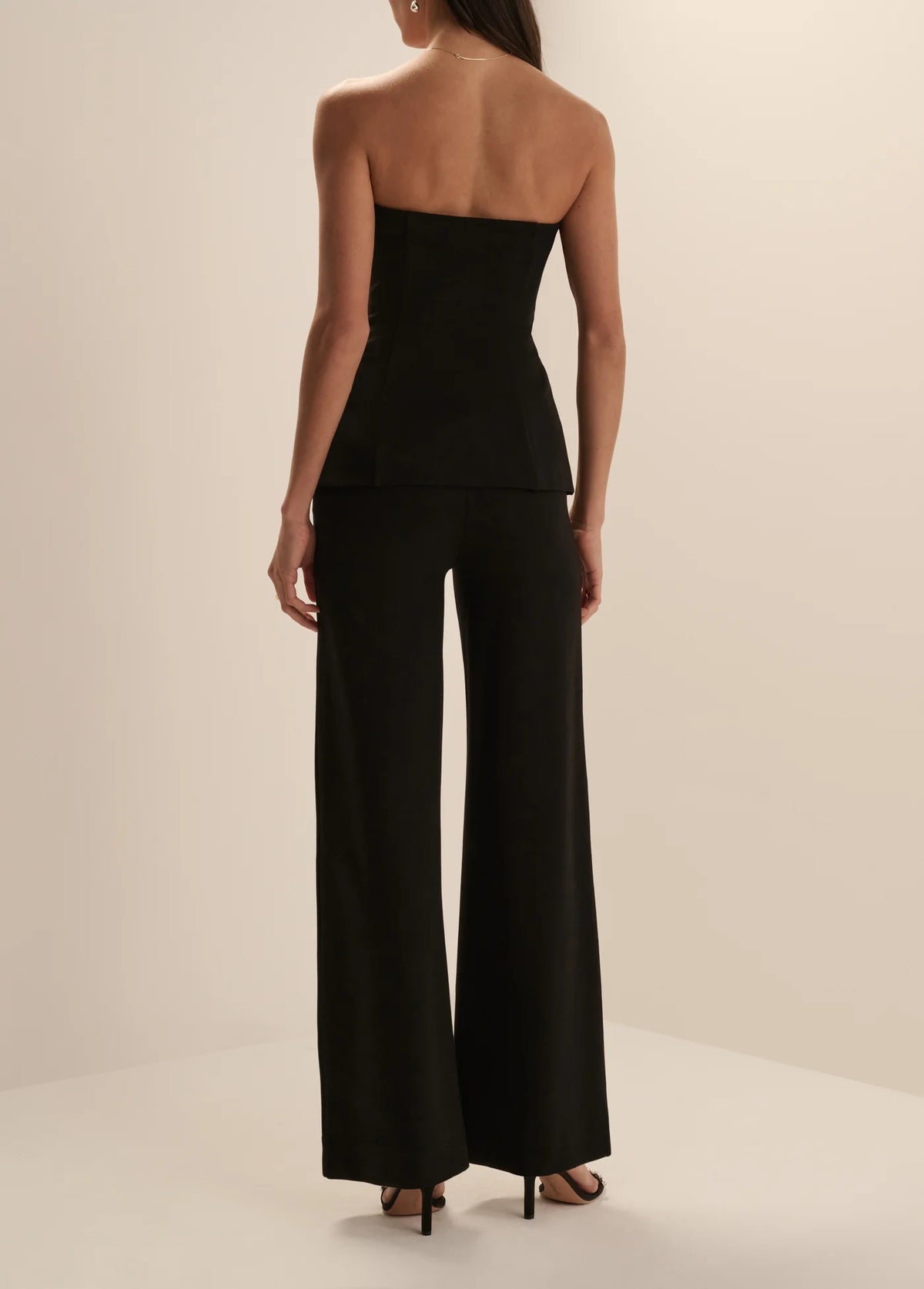 A woman modeling a black strapless top and matching wide-leg trousers set, paired with black heeled sandals, in a minimalist cream studio setting.