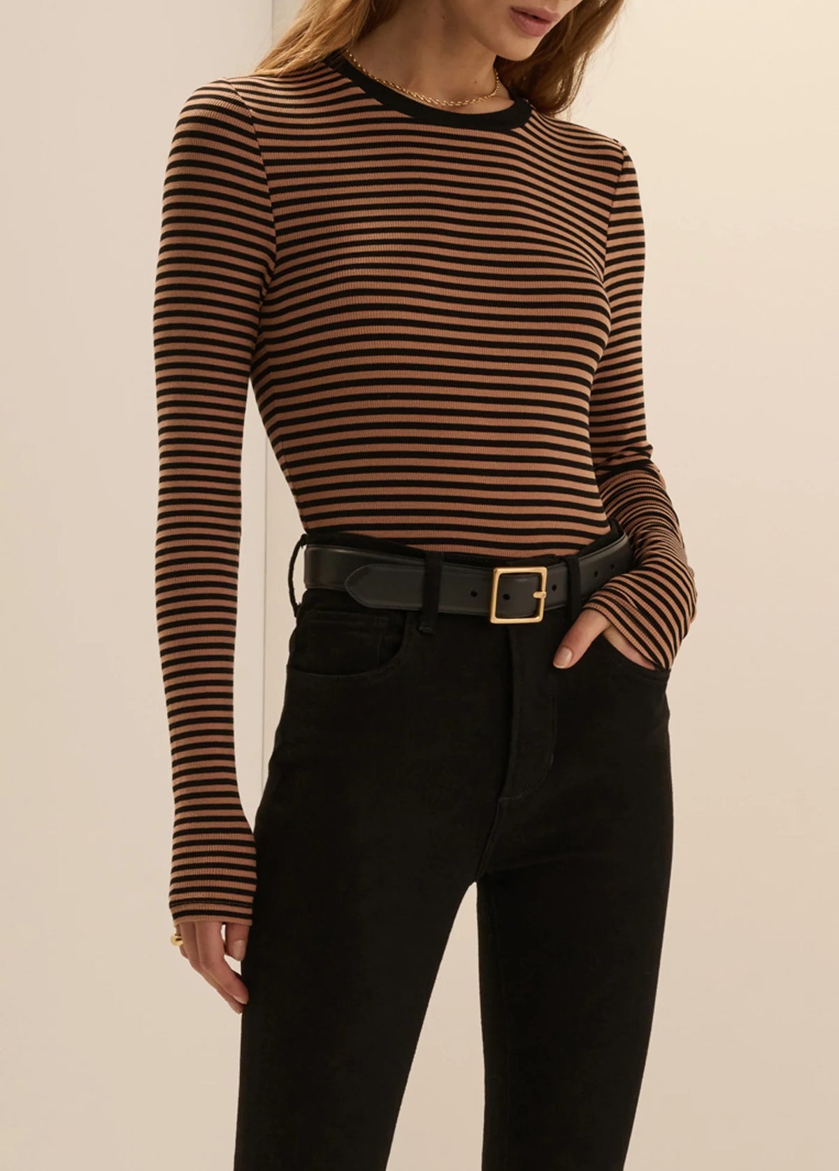A woman wearing a fitted long-sleeve top with thin brown and black horizontal stripes, paired with black pants and a black belt with a gold buckle, against a plain beige studio background.