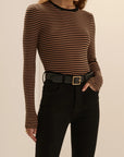 A woman wearing a fitted long-sleeve top with thin brown and black horizontal stripes, paired with black pants and a black belt with a gold buckle, against a plain beige studio background.