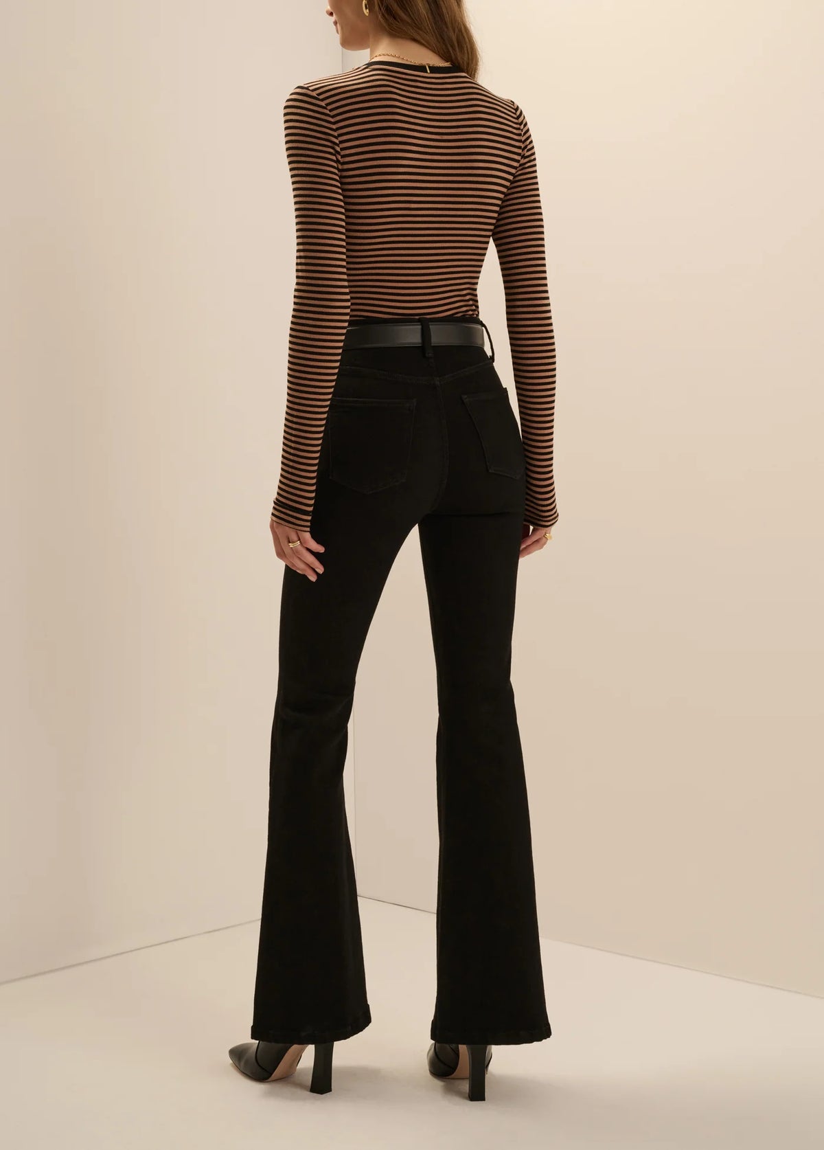 A model shown from the back wearing a brown and black striped long-sleeve mock neck top, black flared jeans, and black heels, set against a plain beige studio background.