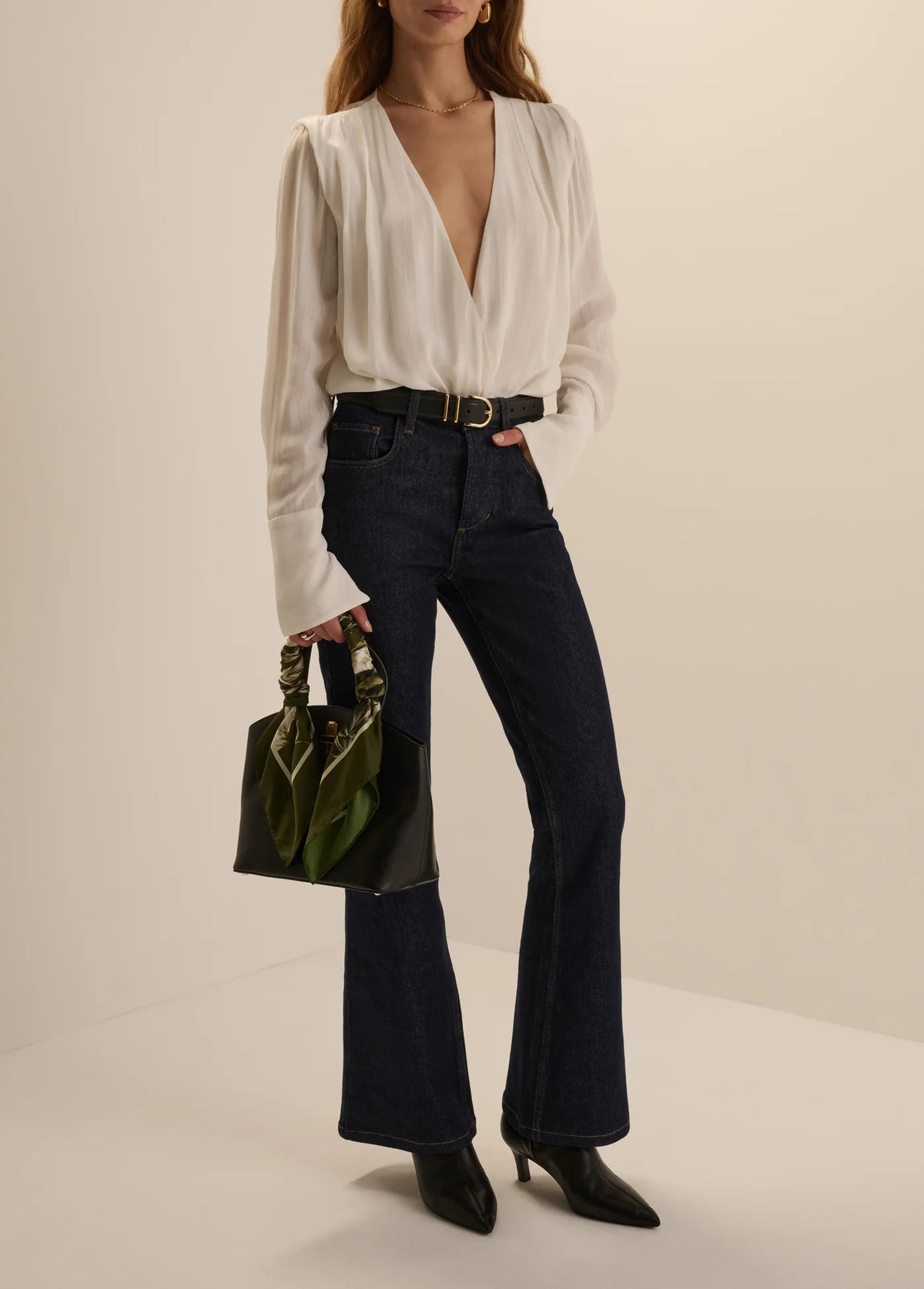 A model wearing a white long-sleeve blouse with a deep V-neck, dark blue high-waisted flare jeans, and black pointed-toe heeled boots, holding a green handbag against a warm beige studio background.