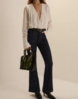A model wearing a white long-sleeve blouse with a deep V-neck, dark blue high-waisted flare jeans, and black pointed-toe heeled boots, holding a green handbag against a warm beige studio background.