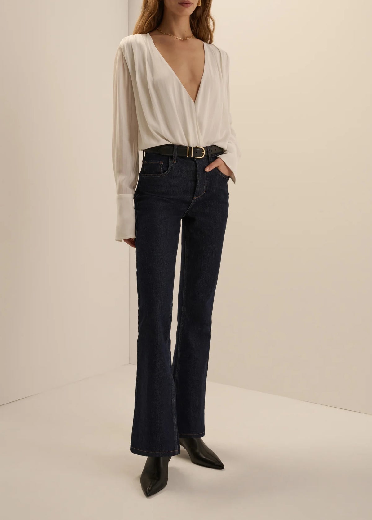 A model wearing dark blue flare jeans with a black belt, paired with a white long-sleeve blouse and black pointed-toe boots, standing against a plain beige studio background.