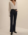 A model wearing dark blue flare jeans with a black belt, paired with a white long-sleeve blouse and black pointed-toe boots, standing against a plain beige studio background.