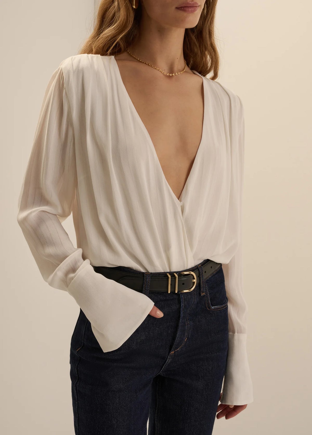 A woman wearing a cream-colored long-sleeve wrap blouse with a deep V-neckline and flared cuffs, paired with dark blue jeans and a black belt with a gold buckle, against a plain beige background.