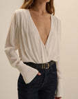 A woman wearing a cream-colored long-sleeve wrap blouse with a deep V-neckline and flared cuffs, paired with dark blue jeans and a black belt with a gold buckle, against a plain beige background.