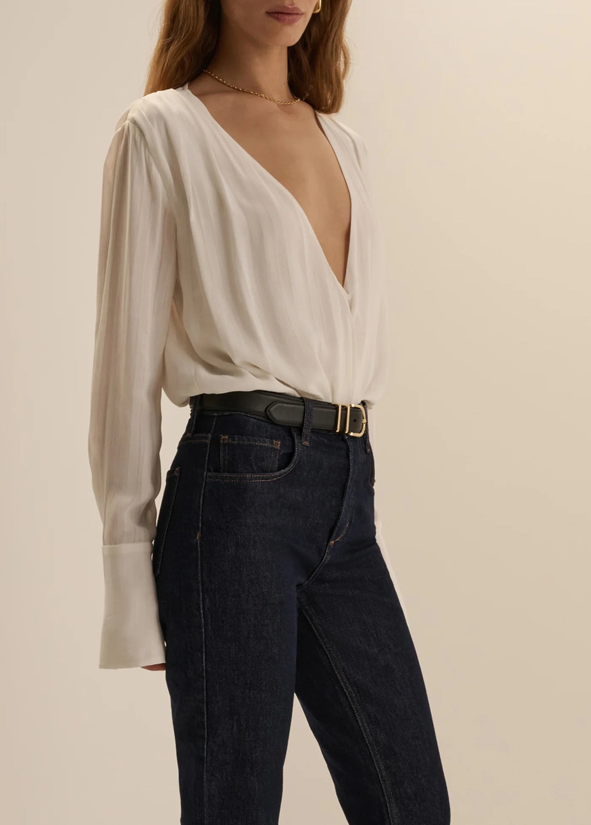 A woman wearing a white long-sleeve blouse with a deep V-neck and dark blue jeans, photographed against a plain beige studio background.