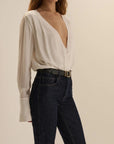 A woman wearing a white long-sleeve blouse with a deep V-neck and dark blue jeans, photographed against a plain beige studio background.
