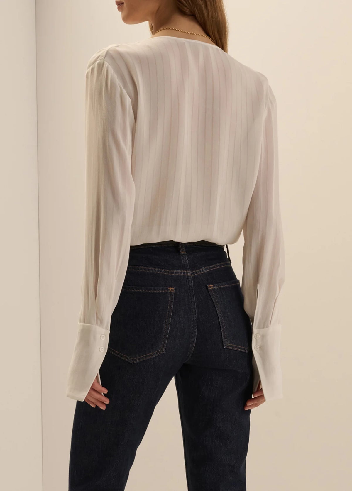 A woman shown from the back wearing a cream-colored, long-sleeved pleated blouse tucked into dark wash jeans.
