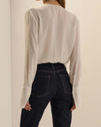 A woman shown from the back wearing a cream-colored, long-sleeved pleated blouse tucked into dark wash jeans.