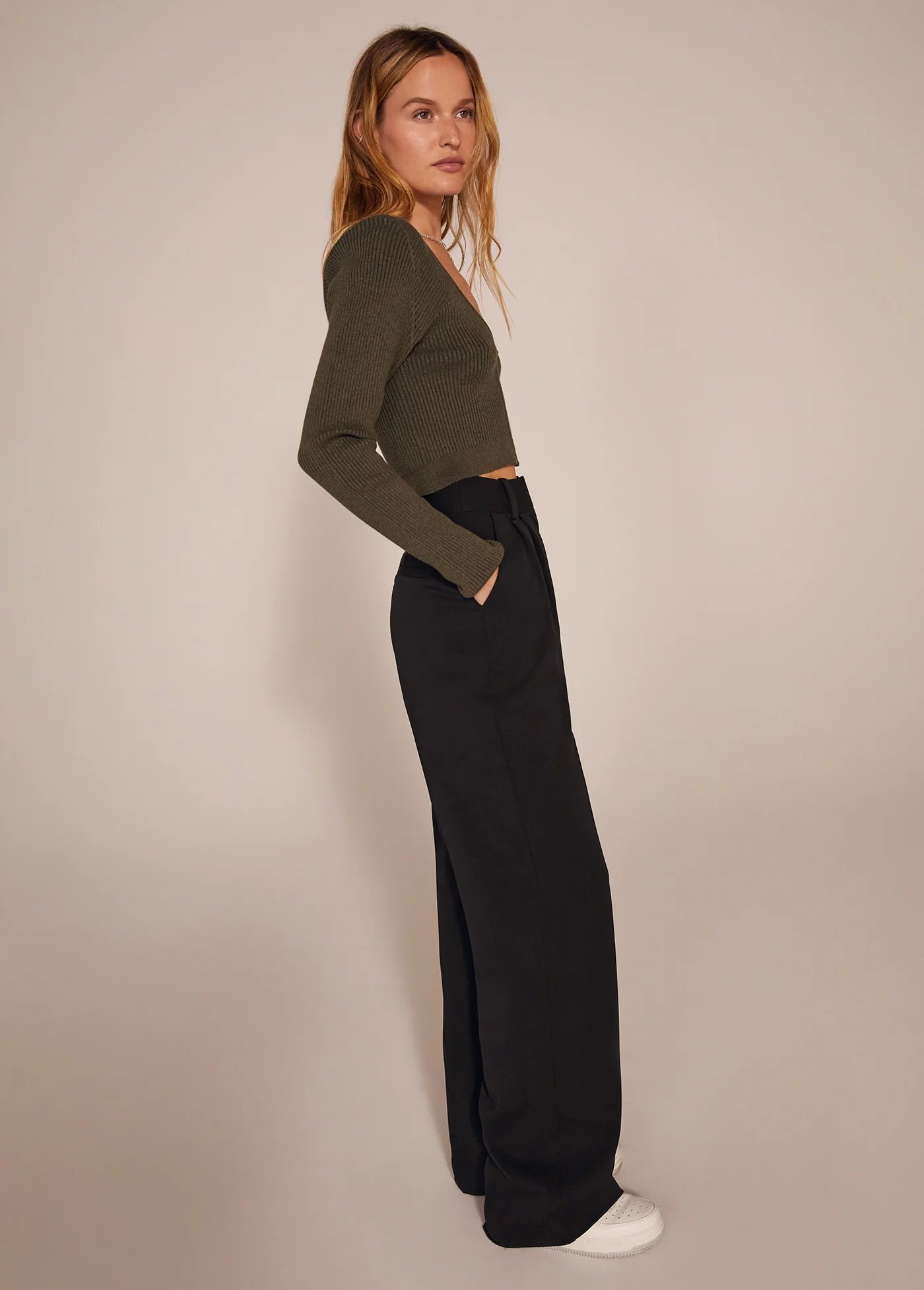 Woman wearing a green sweater and black pants against a beige background, side view