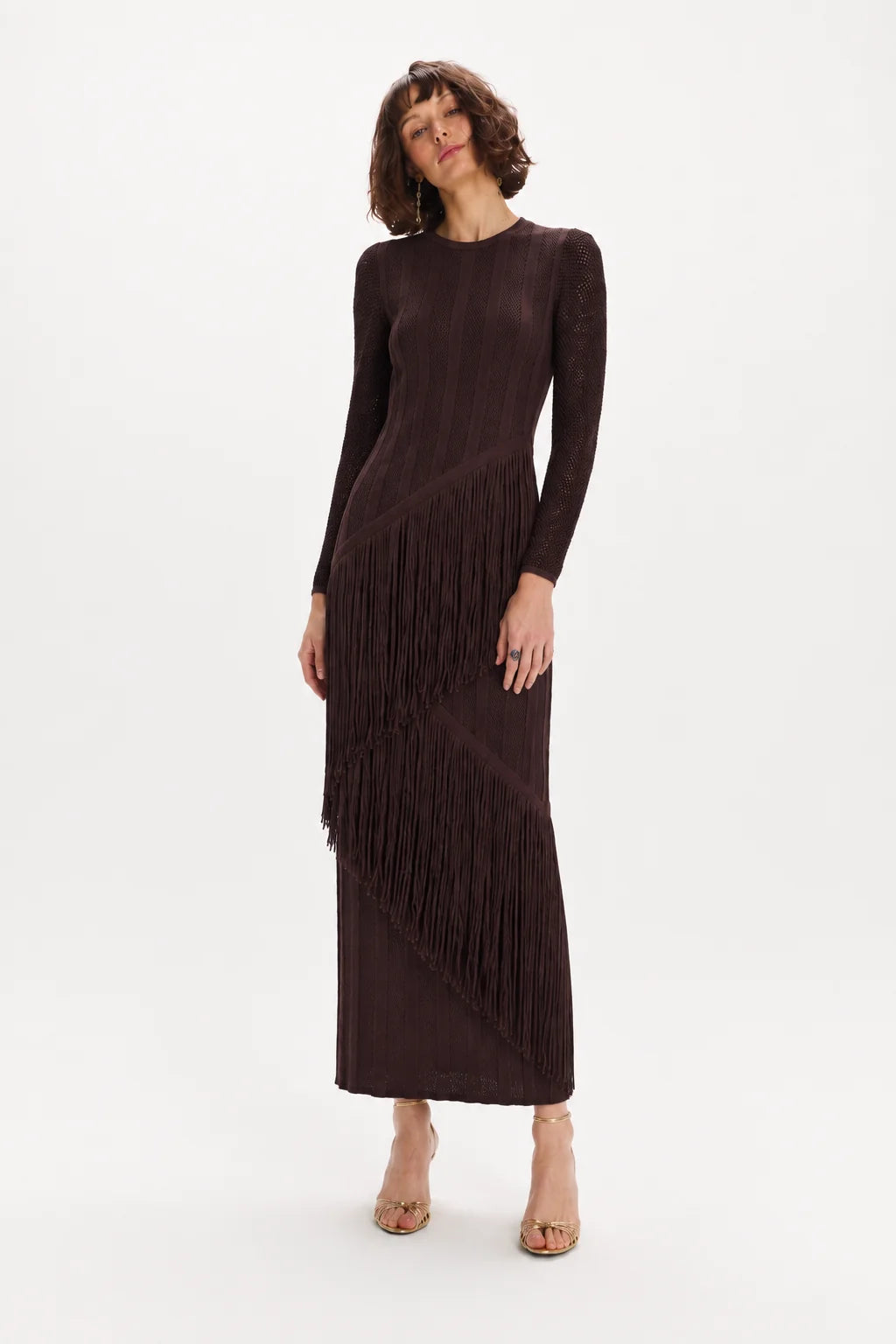 A woman modeling a long-sleeve, dark brown fringe maxi dress against a plain white studio background.