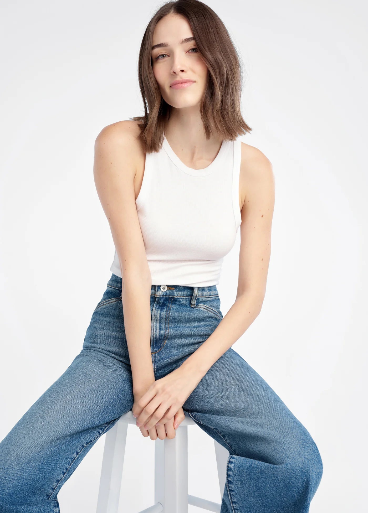 Woman wearing a white tank top and blue jeans sitting on a white stool against a white background