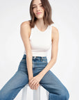 Woman wearing a white tank top and blue jeans sitting on a white stool against a white background