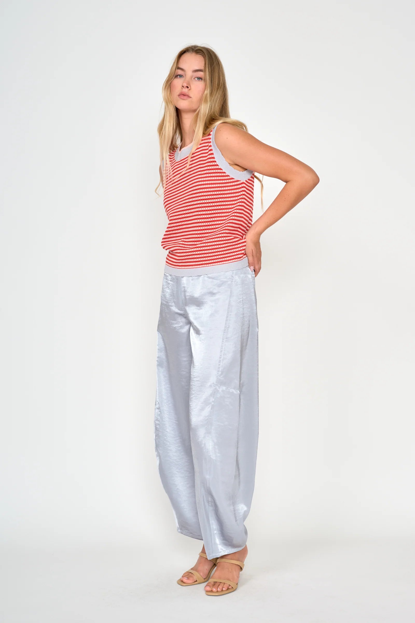 A woman modeling a sleeveless red and white striped top paired with shiny light silver wide-leg pants and beige sandals, standing against a plain white studio background.