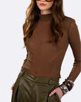 A woman wearing a fitted brown mock-neck long-sleeve top and olive green faux-leather pants, styled with large tortoiseshell sunglasses and chunky metal bracelets against a light gray studio background.