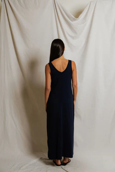 Woman wearing a long dark blue dress standing against a plain white curtain.