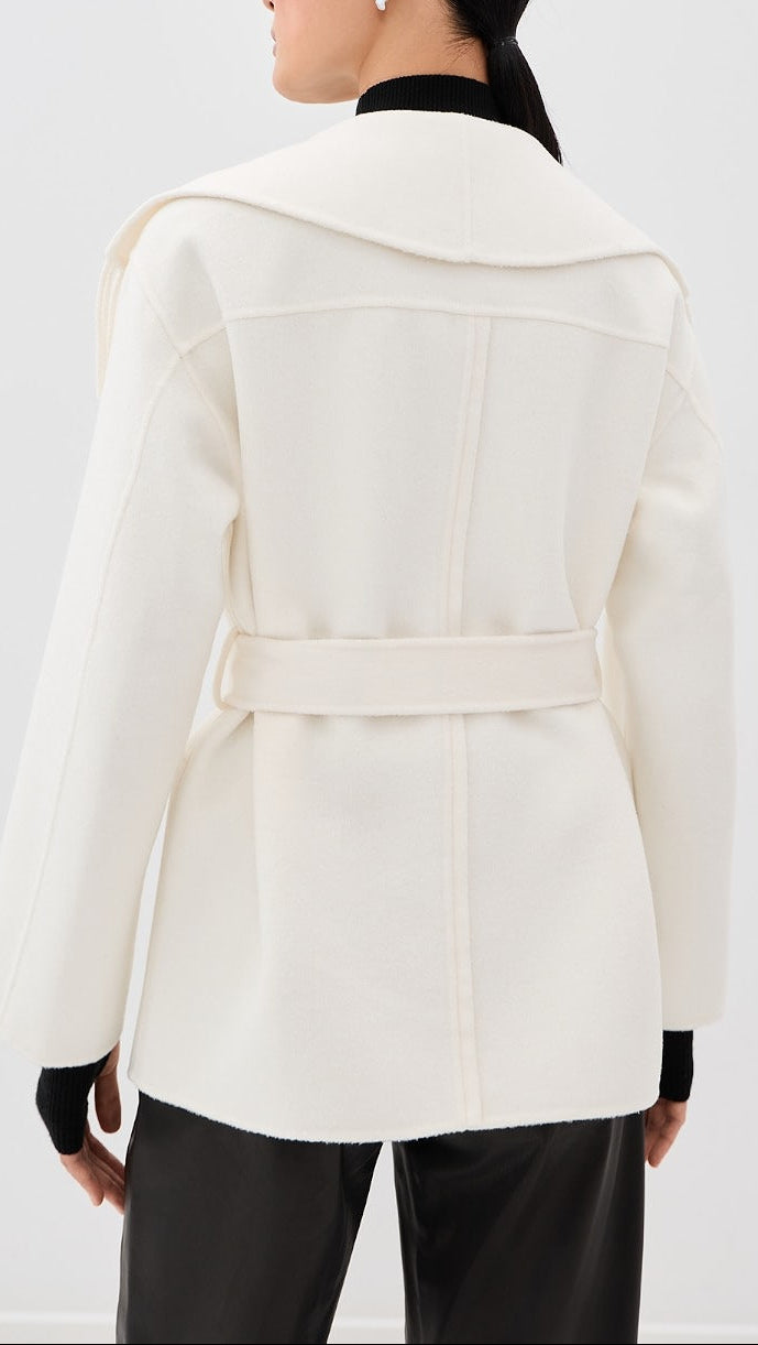 A woman shown from the back wearing a structured cream-colored coat with a wide collar and waist tie, paired with black pants and a black top underneath.