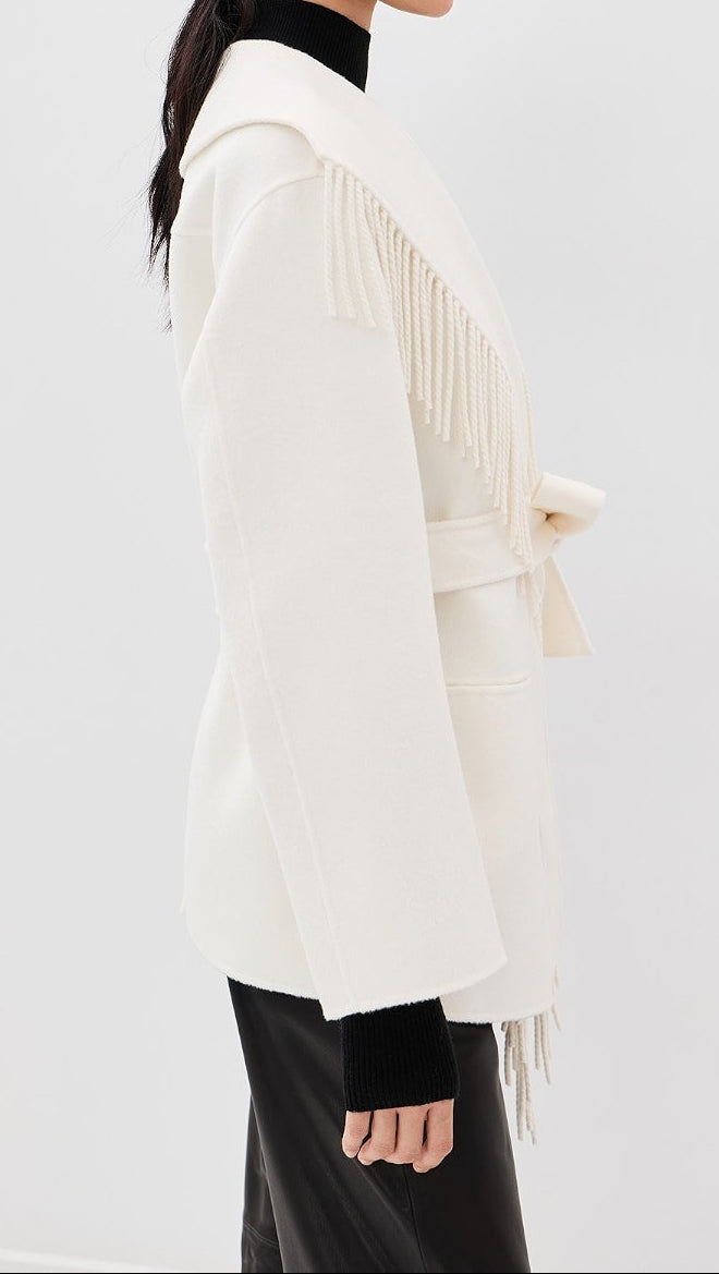 A woman shown in side profile wearing a cream-colored wrap coat with fringe detailing and a tie belt, layered over a black turtleneck and black pants, against a plain light gray studio background.