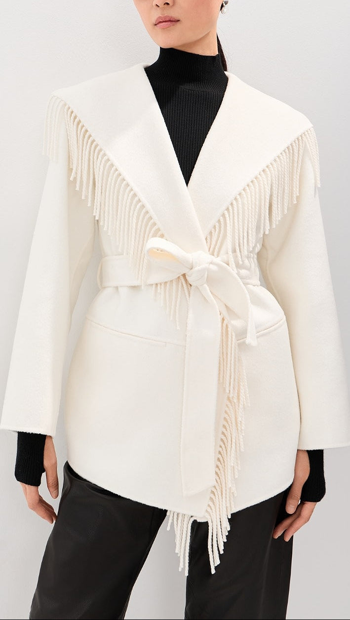 A model wearing a cream-colored wrap jacket with fringe detailing and a tie belt, styled over a black turtleneck and black pants against a light gray background.