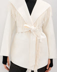 A model wearing a cream-colored wrap jacket with fringe detailing and a tie belt, styled over a black turtleneck and black pants against a light gray background.