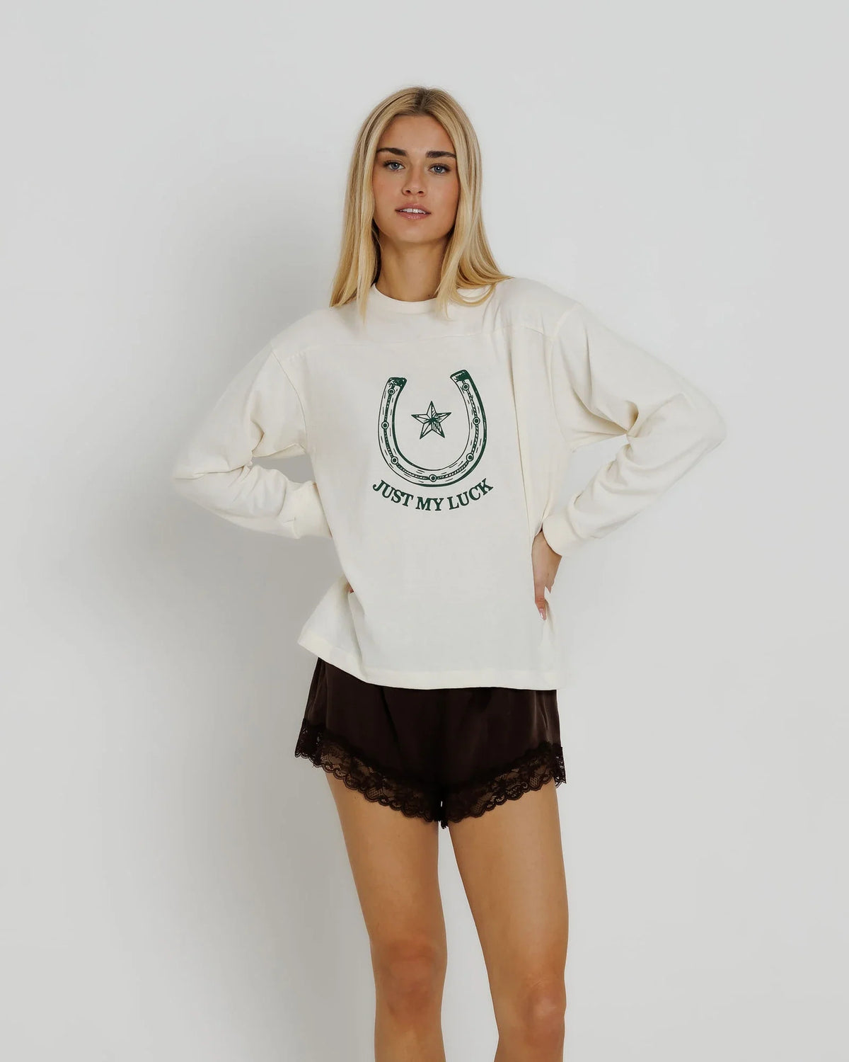 A blonde woman modeling a cream-colored long-sleeve sweatshirt with a green horseshoe graphic and the text 'JUST MY LUCK' on the front, paired with dark brown lace-trimmed shorts, against a light gray studio background.