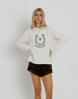 A blonde woman modeling a cream-colored long-sleeve sweatshirt with a green horseshoe graphic and the text 'JUST MY LUCK' on the front, paired with dark brown lace-trimmed shorts, against a light gray studio background.