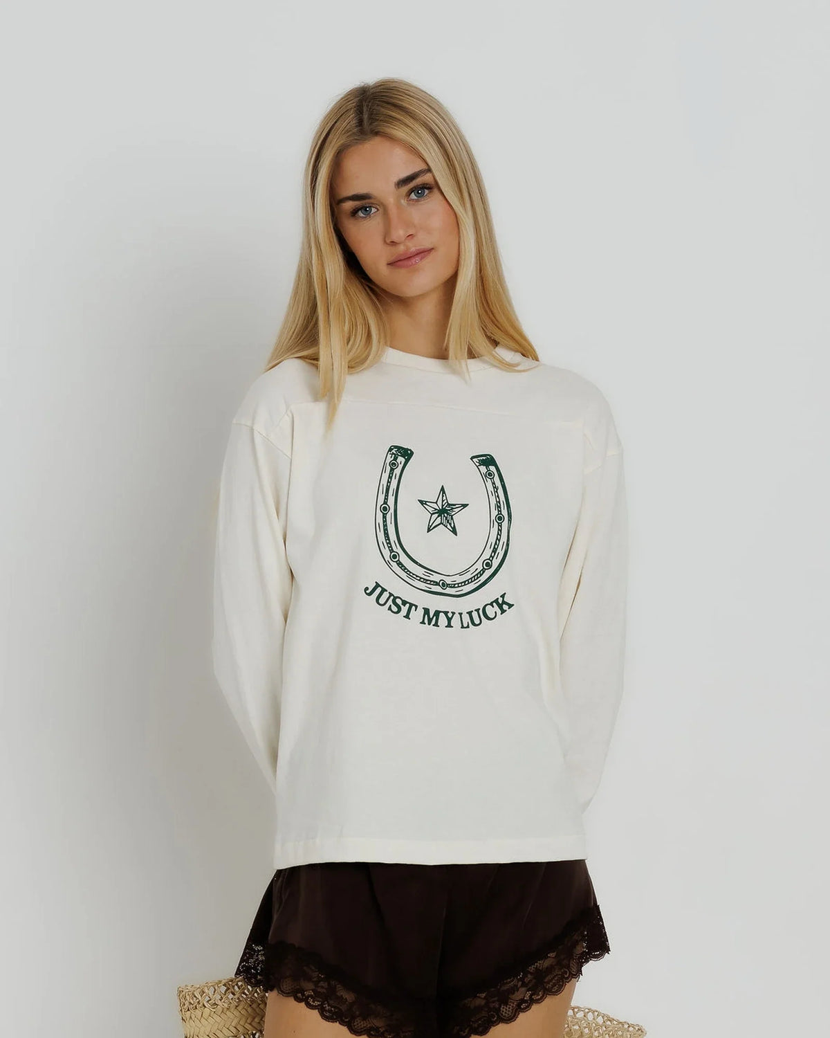 A blonde woman wearing a cream-colored long-sleeve sweatshirt with a dark green horseshoe and star graphic and the text 'JUST MYLUCK' on the front, paired with dark brown lace-trimmed shorts, against a plain light gray background.