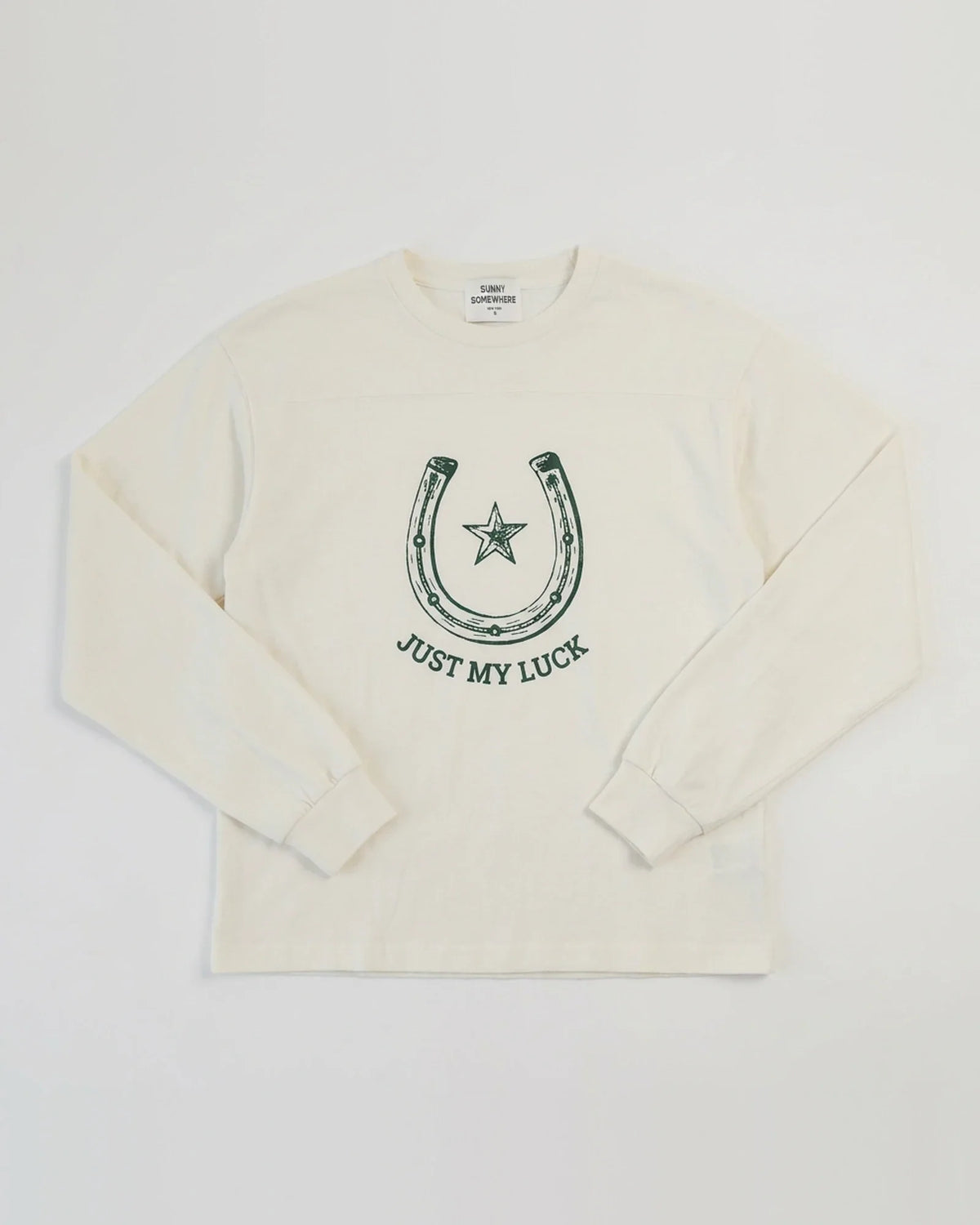 An off-white long-sleeve crewneck sweatshirt featuring a dark green graphic of a horseshoe with a star and the text 'JUST MY LUCK' printed on the chest.