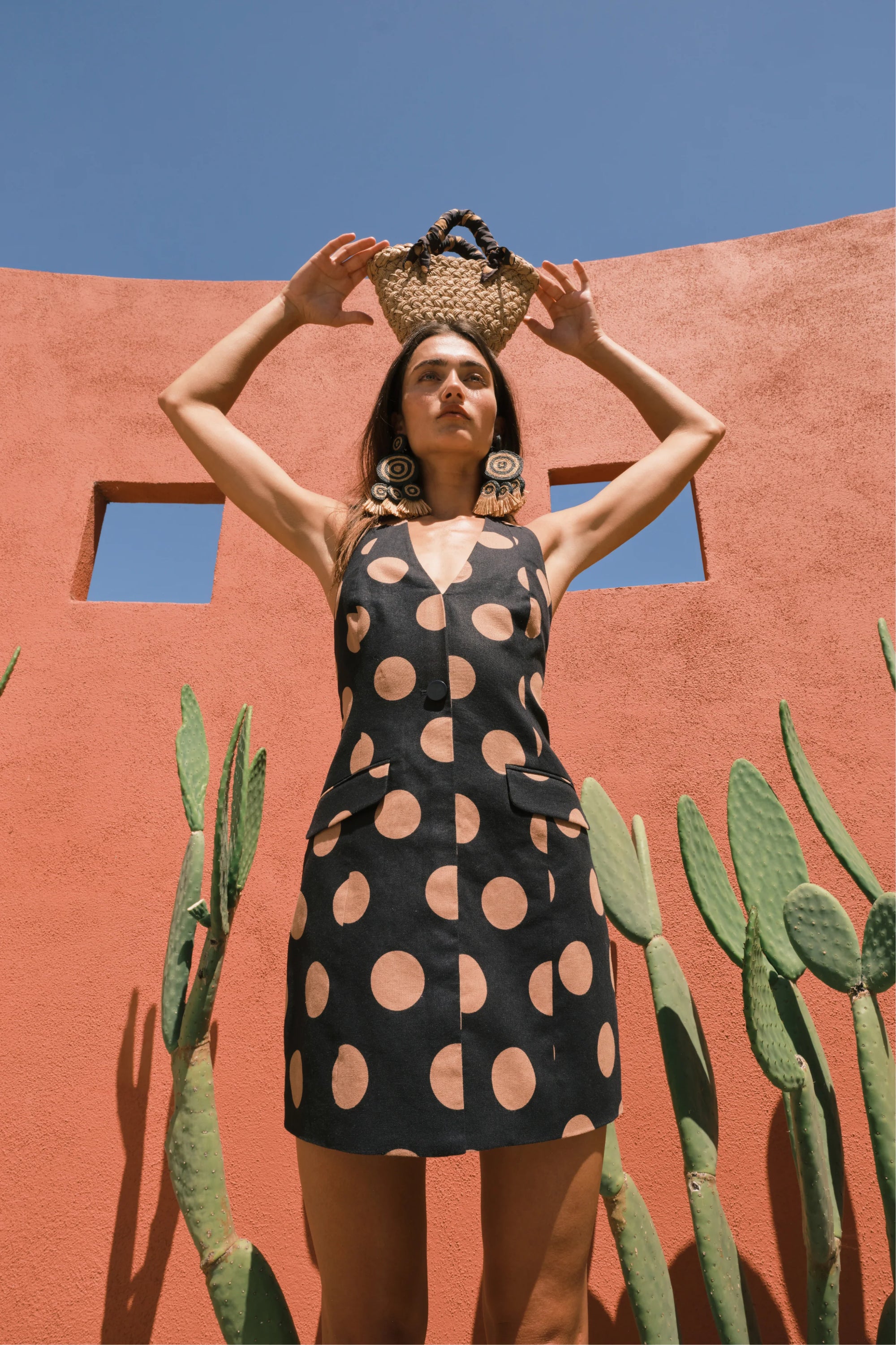 Woman in a black dress with tan polka dots holding a woven basket against a red wall with cacti.
