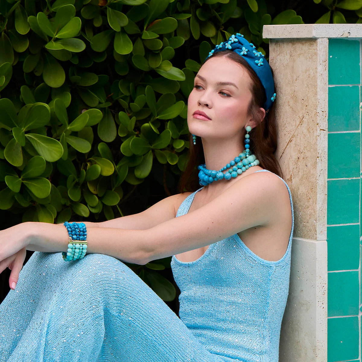 Woman in a blue dress and accessories sitting against a green tiled wall.