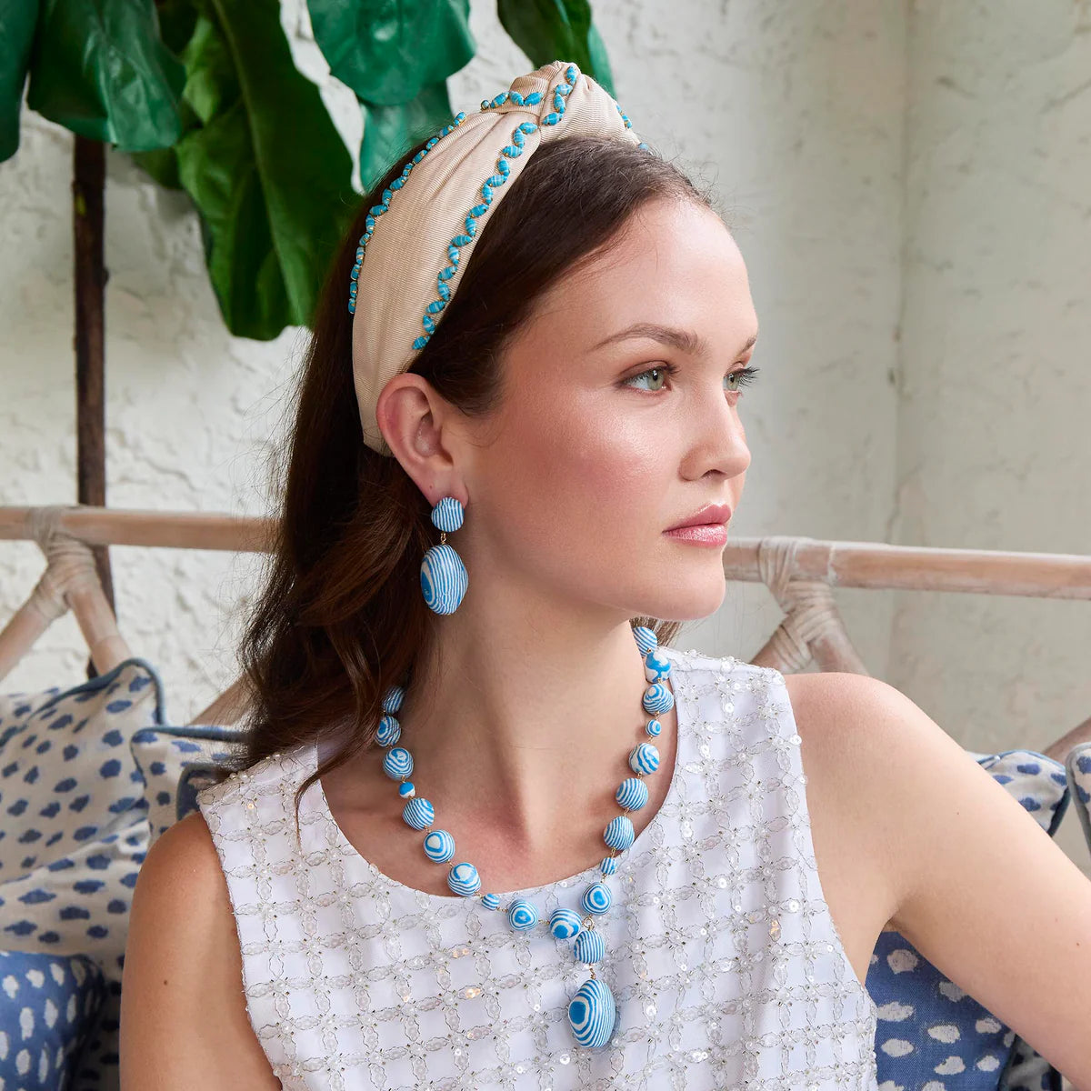 model with a blue and white necklace and earrings with a headband sitting on a couch with a plant in the background 
