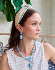 model with a blue and white necklace and earrings with a headband sitting on a couch with a plant in the background 