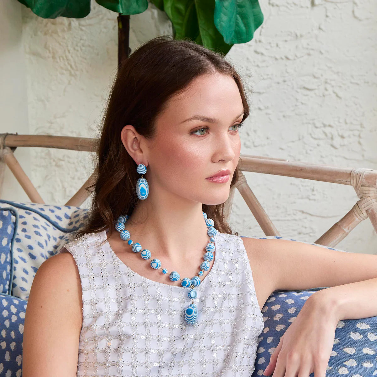 model with blue and white jewelry sitting with a couch and plant in the background