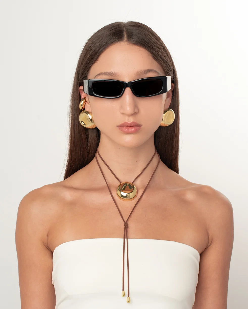 A model with long dark hair is shown from the chest up, wearing a white strapless top, gold hoop earrings, a brown cord necklace with a gold pendant, and black cat-eye sunglasses against a plain white background.