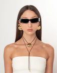 A model with long dark hair is shown from the chest up, wearing a white strapless top, gold hoop earrings, a brown cord necklace with a gold pendant, and black cat-eye sunglasses against a plain white background.