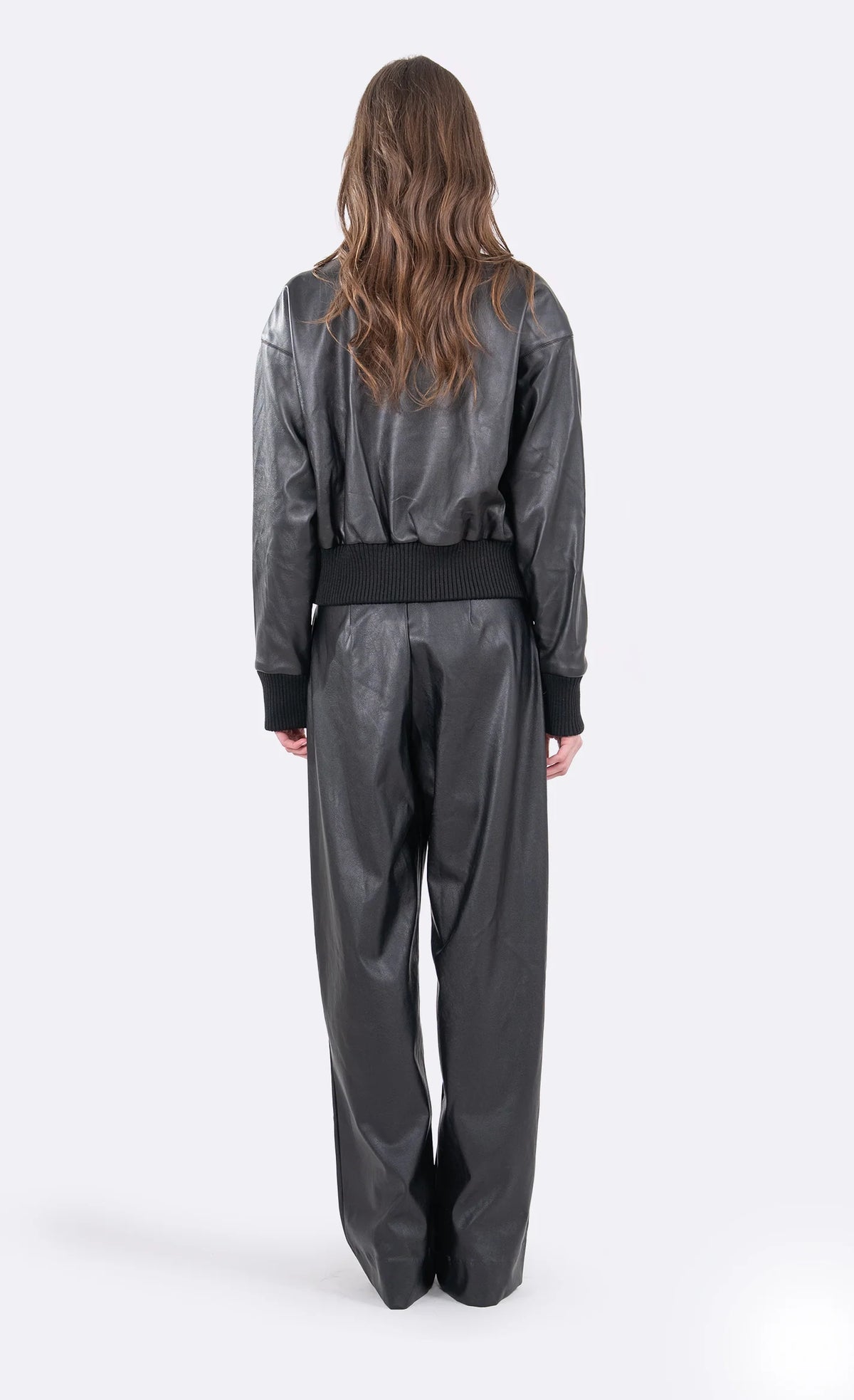 A model viewed from the back wearing a black faux-leather jumpsuit with wide-leg pants, long sleeves, and a matching black waist belt, set against a plain light gray studio background.