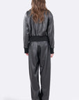 A model viewed from the back wearing a black faux-leather jumpsuit with wide-leg pants, long sleeves, and a matching black waist belt, set against a plain light gray studio background.