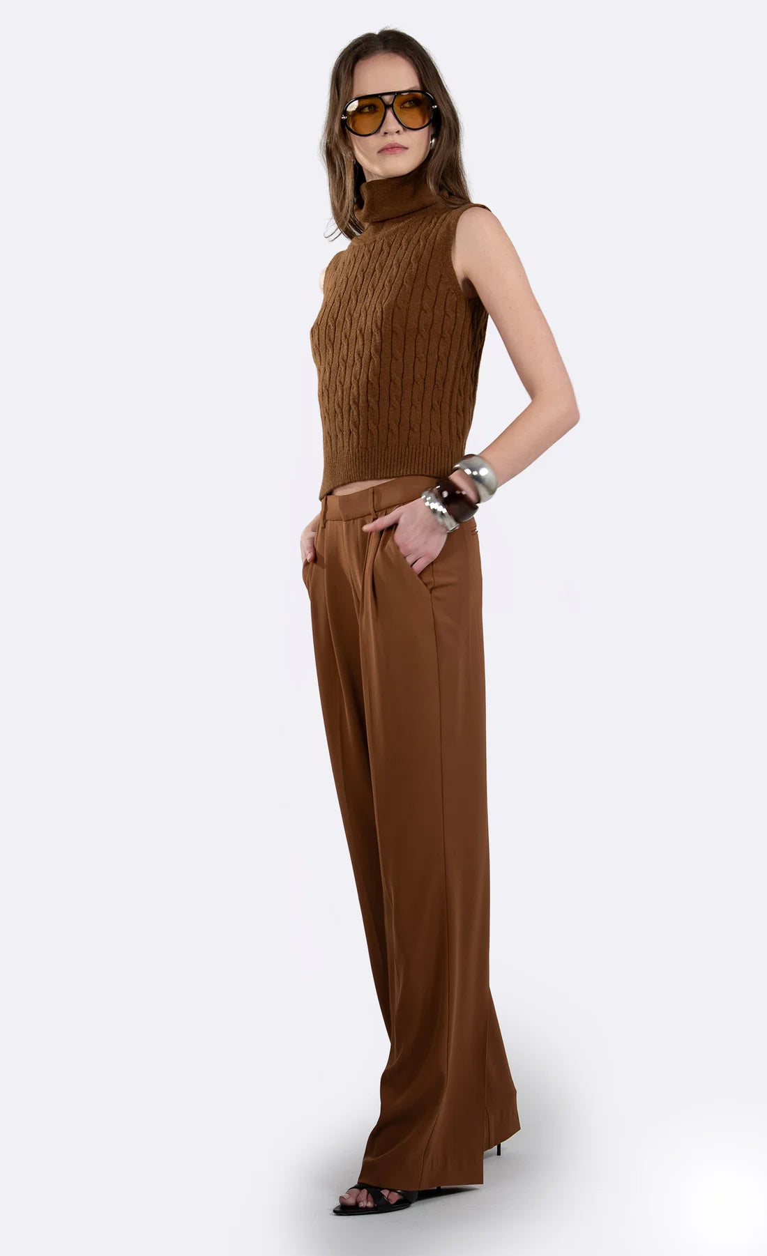 A woman modeling a brown sleeveless mock-neck knit top paired with matching wide-leg trousers, styled with sunglasses and bracelets against a light gray background.
