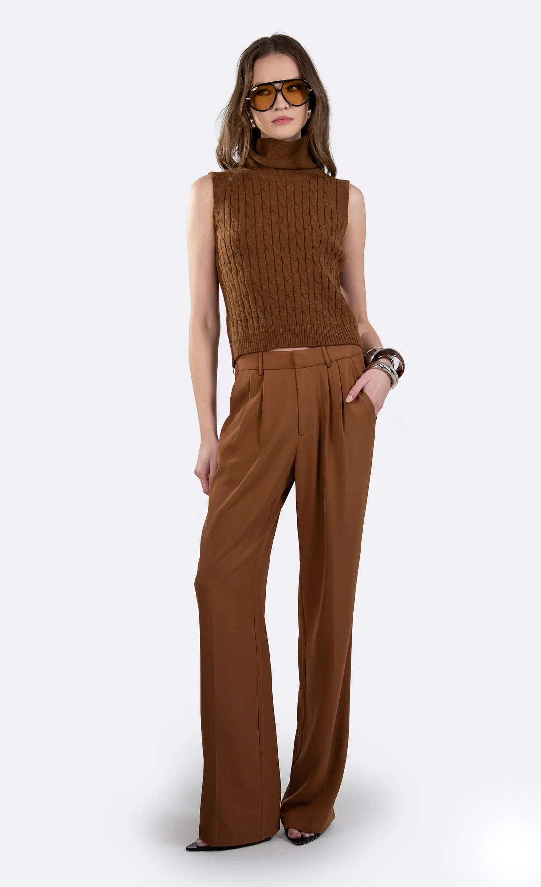 A woman modeling a brown sleeveless ribbed knit mock-neck top paired with matching brown wide-leg trousers, styled with tortoiseshell sunglasses and bracelets against a light gray studio background. full body front