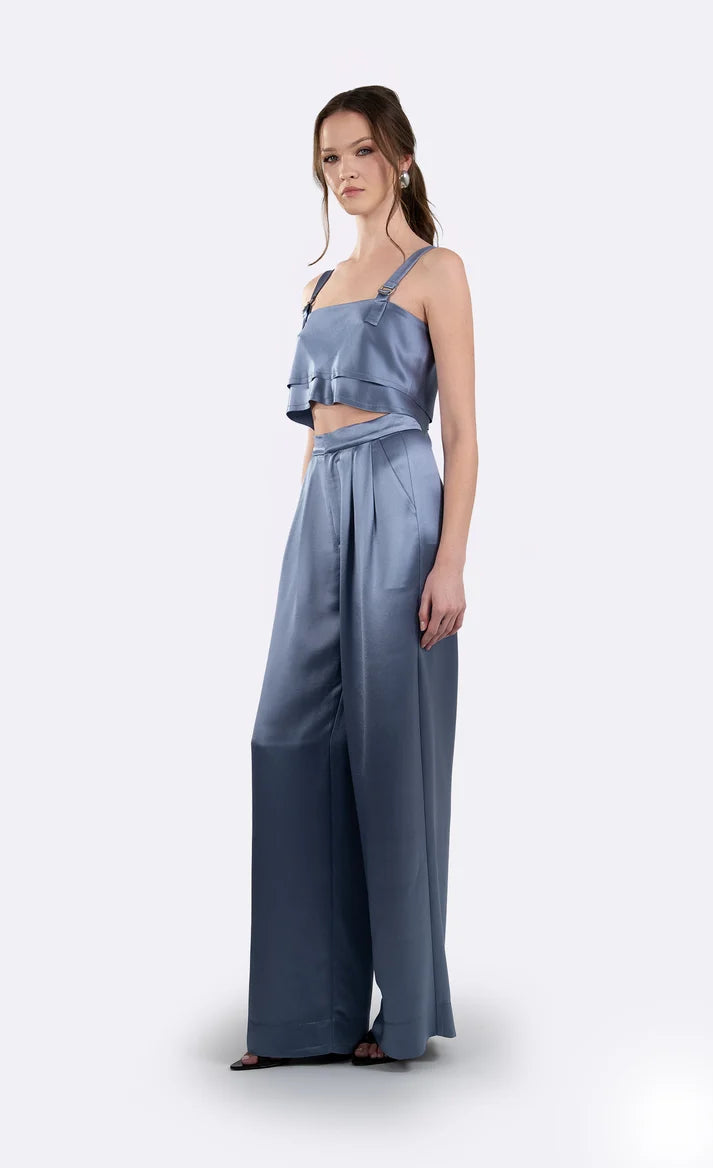 A woman modeling a two-piece satin outfit consisting of a light blue ruffled crop top and matching wide-leg pants in a dusty blue shade, set against a plain light gray background. side/front view