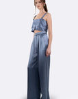 A woman modeling a two-piece satin outfit consisting of a light blue ruffled crop top and matching wide-leg pants in a dusty blue shade, set against a plain light gray background. side/front view