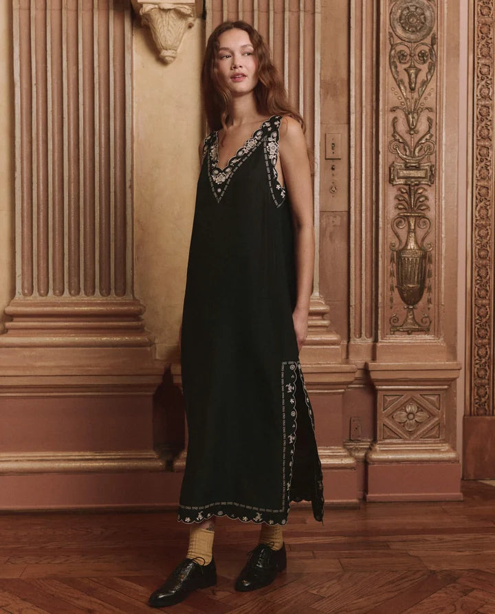 A woman stands in an ornate interior wearing a long black dress with silver embroidered detailing around the neckline and hem, paired with beige socks and black shoes.
