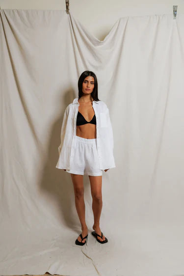 Woman wearing a white shirt and shorts with a black bralette against a plain background