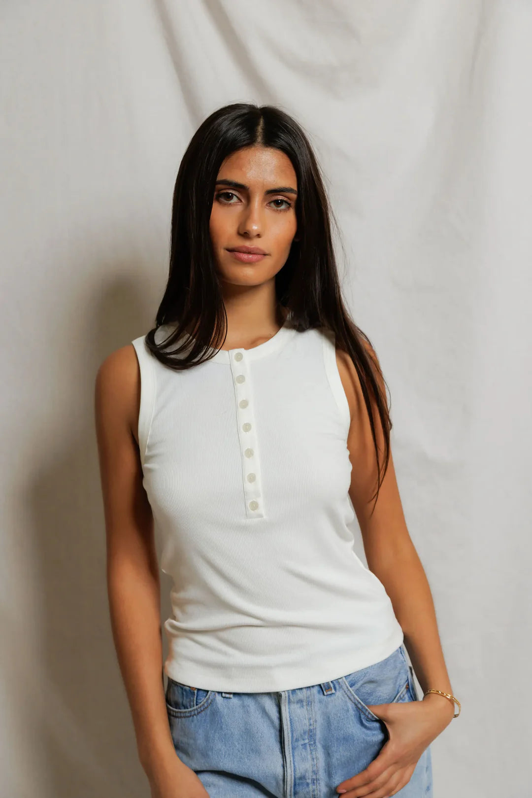 Woman wearing a white sleeveless top and blue jeans against a plain background